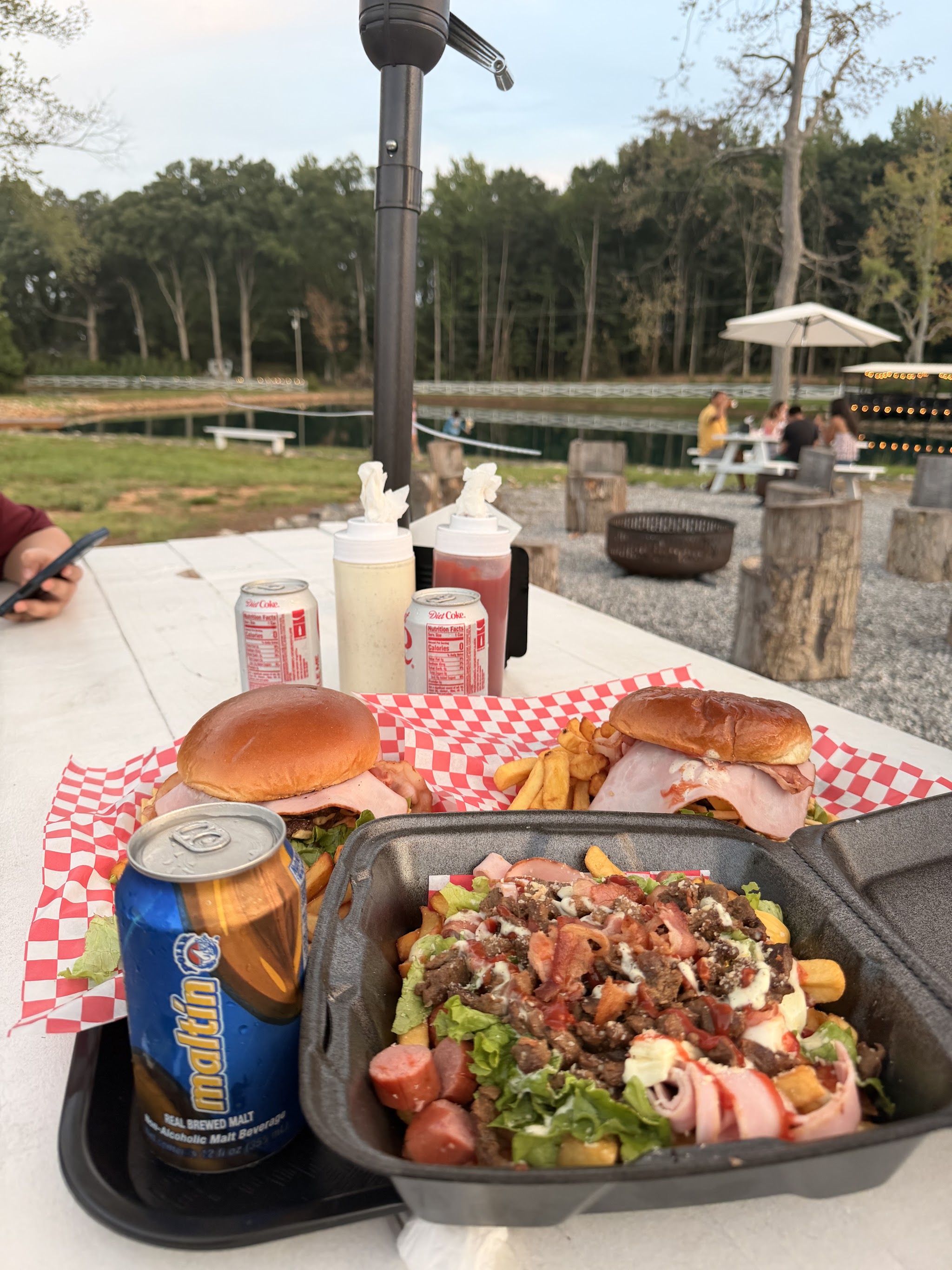 Burgers, a loaded fries box and a maltín on a picnic table beside the lake